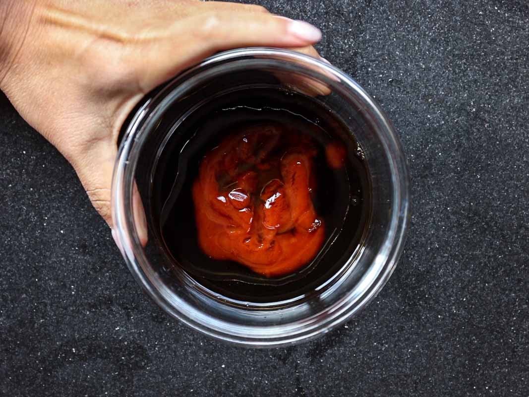A hand holds a clear glass against a dark surface. Inside the glass is a dark liquid with a swirl of reddish sauce on top.